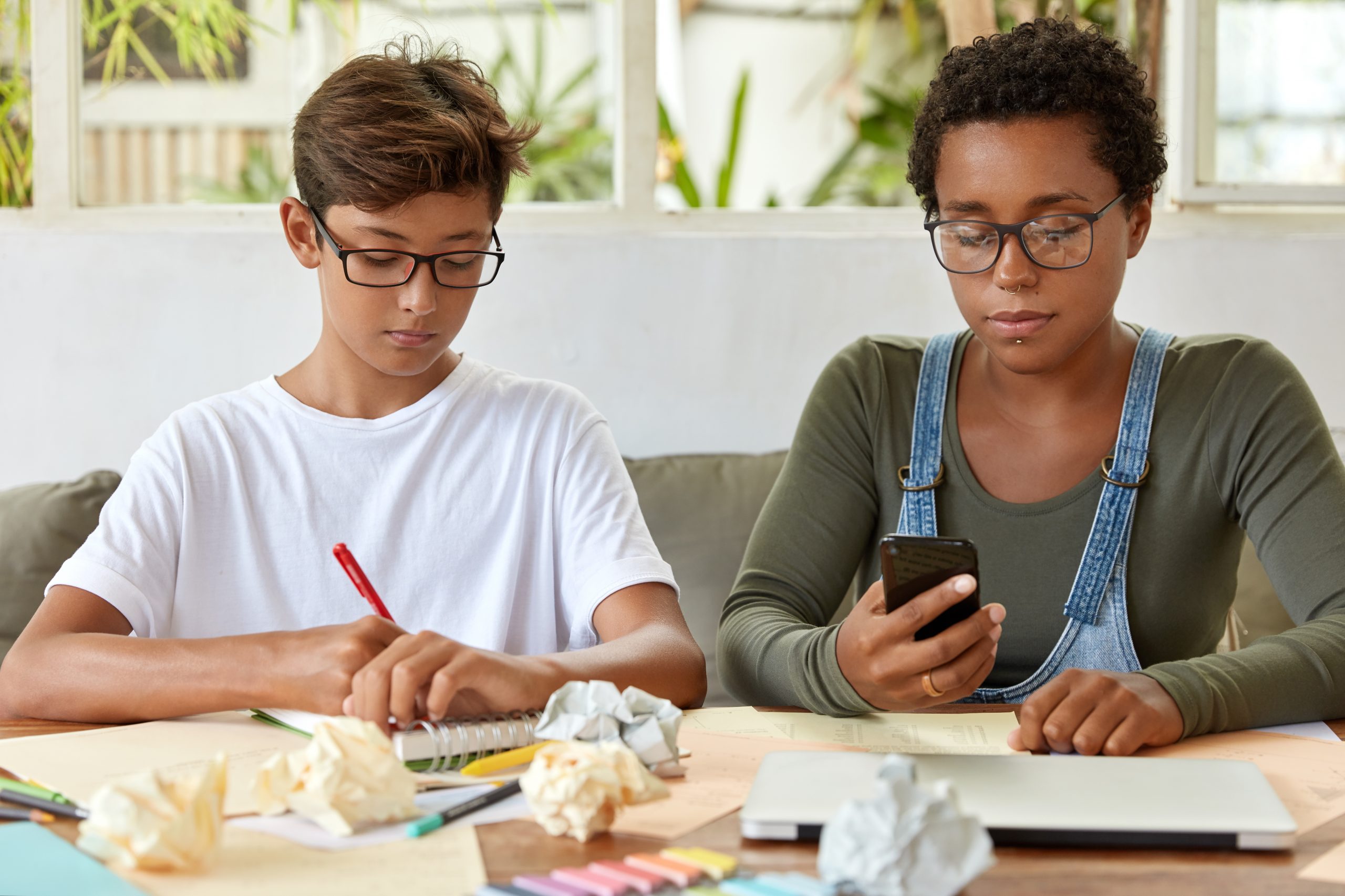 Handsome youngster in casual t shirt, writes down instruction in organizer, his black female friend uses smart phone, connected to wireless internet. Two mixed race boy and girl collaborate together